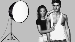 Two models pose together in a studio with a large softbox light positioned to the left.