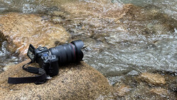 Professional DSLR camera with telephoto lens resting on a rock beside flowing water.