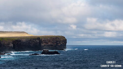 Dramatic coastal cliffs with turquoise waves crashing against dark basalt rock formations under a partly cloudy sky.