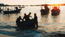 Silhouetted fishermen wade in shallow water carrying a net at golden hour with fishing boats anchored in the background.