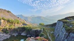 Alpine valley landscape with layered mountains, rocky cliffs, and winding river below clear sky.