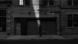 Solitary figure in dark clothing standing against black storefront on brick building facade in monochrome light.