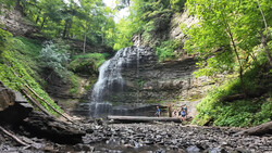 Cascading waterfall flowing over layered rock formations in a lush forested gorge.