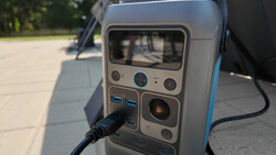 Portable power station with display screen, control buttons, and USB ports, photographed outdoors on pavement.