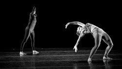 Two dancers in dramatic poses on a dark stage, captured in high-contrast black and white lighting.