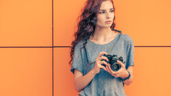 Woman with long wavy hair holding a vintage film camera against an orange background.