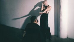 Shirtless man against white wall with dramatic shadow of another person cast across his torso.