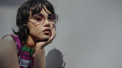 Young person wearing clear-framed glasses and colorful striped tank top, hand to face in contemplative pose against neutral background.