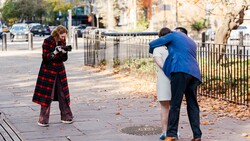 Man in blue blazer posing while woman in red plaid coat photographs him on an urban sidewalk.
