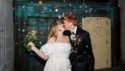 Newlyweds kissing in front of a teal doorway decorated with hanging paper lanterns and confetti.