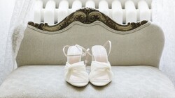 Pair of white infant shoes displayed on a cream-colored vintage chair with ornate wooden frame.