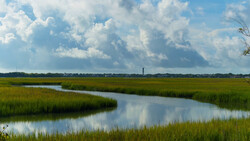 Winding waterway curves through expansive marsh landscape under cumulus clouds.