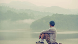 Person sitting alone on a rocky shore, gazing across a misty lake toward forested mountains.