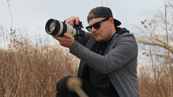 Photographer holding a telephoto lens camera while looking through the viewfinder in an open field.