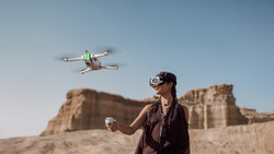Operator wearing VR headset controls a small quadcopter drone in flight above desert terrain.