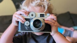 Young child holding a vintage rangefinder camera up to their face, obscuring their head.