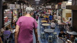 Woman in purple shirt walking through crowded indoor market or fair with vendor booths and tables.