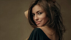 Woman with long dark hair looking over shoulder against neutral tan background.