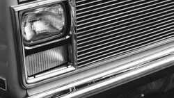 Close-up black and white photograph of a classic car's front end, showing rectangular headlight and horizontal grille detail.