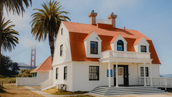 White Victorian-style house with distinctive orange-red mansard roof and dormer windows, palm trees nearby, suspension bridge visible in background.