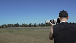 Photographer from behind holding a telephoto lens, shooting across an open field under clear blue sky.