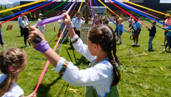 Children holding colored ribbons around a maypole on a grass field during a community event.