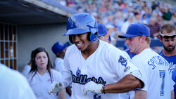 Baseball player in blue uniform smiling while being congratulated by teammates in dugout.