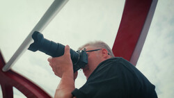 Photographer holding telephoto lens up to eye while shooting outdoors against cloudy sky.