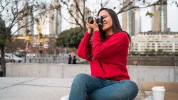 Woman in red jacket holding binoculars to her eyes while sitting outdoors near urban architecture.