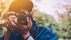 Photographer holding a DSLR camera up to their face, composing a shot outdoors in natural sunlight.
