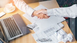 Two people in white shirts shake hands over a desk during a business meeting.