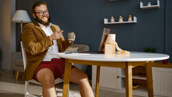 Bearded man in mustard blazer and plaid shorts sits at round table with laptop and coffee cup.