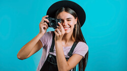 Young woman holding vintage film camera up to her face, wearing black hat and overalls against bright turquoise background.