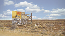 Weathered vintage billboard with faded yellow and red paint stands alone in a sparse desert landscape under blue sky with white clouds.