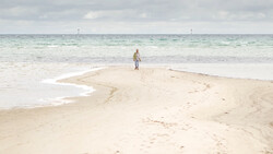 Lone figure standing on a curved sandbar extending into shallow turquoise water.