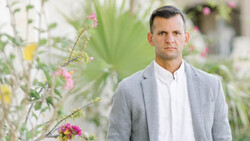 Man in light gray blazer and white shirt standing in front of blooming flowers and foliage.