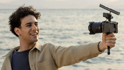 Videographer holding a cinema camera with microphone at a beach location.