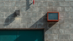 Minimalist urban wall with concrete blocks, red fire alarm box, and geometric shadows from sunlight.