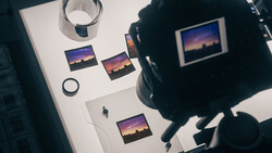 Overhead view of a photographer's workspace with printed sunset landscape photos, a camera, glass of water, and lens cap on white surface.