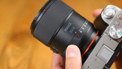 Hand holding a mirrorless camera with a black telephoto lens attached against a warm, blurred orange background.