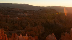 Golden sunrise light illuminates layered hoodoos and forested ridges in a vast canyon landscape.