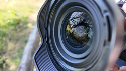 Close-up of a camera lens front element with shallow depth of field, showing tree branches and landscape reflected in the glass.