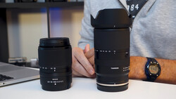 Two black camera lenses displayed side by side on a white surface, with a person's hand holding the larger telephoto lens.