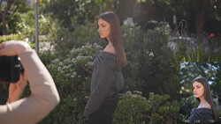 Pregnant woman in dark sweater posing among garden foliage while photographer frames shot in foreground.