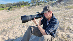 Photographer seated on sand using a telephoto lens while shooting at a beach location.
