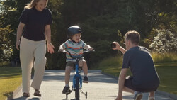 Woman supervises young child riding blue bicycle with training wheels on a paved path as man crouches nearby.