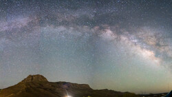 Milky Way galaxy stretches across night sky above a dark mountain silhouette.