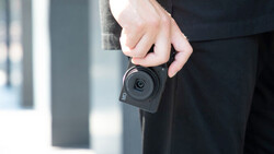 Person in dark clothing holding a compact action camera with a circular lens against their body.