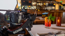Professional video camera on tripod filming colorful cocktails on a white table in a bar setting.
