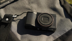 Ricoh GR compact digital camera with textured grip sitting on a gray fabric surface with a wrist strap attached.